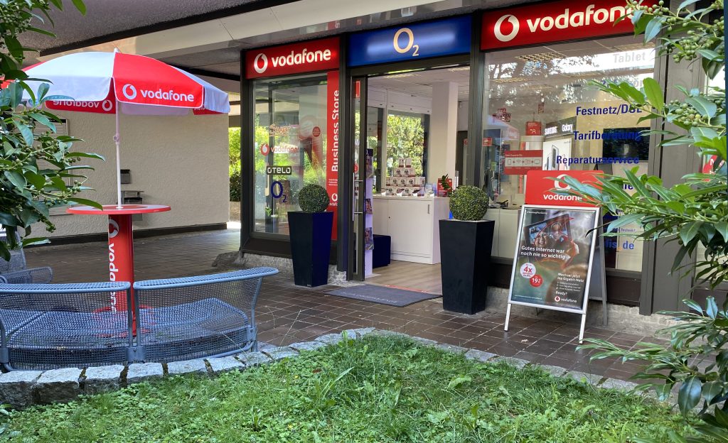 Eingang zu einem Vodafone Shop mit O2 Logo und Außenbereich mit Möbeln.